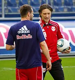 Niko Kovač bei seinem ersten Pflichtspiel als Co-Trainer 2011 von Red Bull Salzburg gekleidet im Trainingsanzug des Vereines mit einem Fußball in der linken Hand im Gespräch mit einer weiteren Person im kurzärmeligen Vereins T-Shirt mit Sponsor und Ausrüster bedruckt zu Kovač gewandt