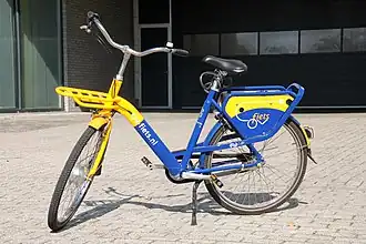Farbfoto. Blau-gelbes E-Hollandrad, tiefer Einstieg, Beleuchtung, Gepäckträger vorne und hinten, Handbremse, Klingel. Auf dem Rahmen steht „OV-fiets.nl“. Städtischer Hintergrund.
