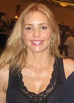 Olivia d'Abo bei der Hollywood Show im Marriott Convention Center in Burbank, Kalifornien, im Jahr 2010. Sie hat lange blonde Haare und trägt ein schwarzes Oberteil mit Spitzenverzierungen am Ausschnitt. Ihr Gesichtsausdruck ist freundlich und offen, und sie scheint sich in einer Gesprächssituation zu befinden. Der Hintergrund ist ein geschäftiges Indoor-Setting mit mehreren Menschen und Ständen.
