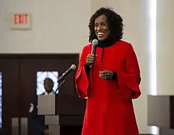 Foto: Eine Frau im roten Mantel spricht in ein Mikrofon, das sie vor ihren Mund hält.