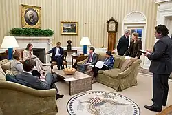 In einer Sitzung mit genütlicher Atmosphäre ist Barack Obama in Gespräch mit Beratern. Die Teilnehmer sitzen auf Sofas und Sessel rund um einen niedrigen Fliesentisch mit einer Obstschale darauf