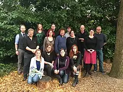 Gruppenbild mit 15 überwiegend fröhlich wirkenden Personen, im Hintergrund Wald