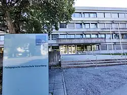 Ein dreistöckiges, graues Gebäude aus Beton mit dem Namen "Pädagogische Hochschule Vorarlberg" buchstabiert in silbernen Buchstaben an der Fassade und einem Schild mit der PH Vorarlberg-Logo im Vordergrund.