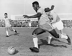 Schwarzweißfotografie vom Spielverlauf zwischen drei Spielern. Zwei Spieler stürmen von rechts nach links dem Fußball hinterher. Im linken Hintergrund ist ein Spieler bereit zum Annehmen.