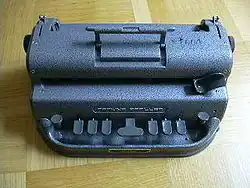 Das Gerät scheint aus Spritzguss zu bestehen, es hat rundliche Konturen und eine blaugraue Hammerschlaglackierung. Vorn sind neun Tasten zu erkennen: in der Mitte die breite Zwischenraumtaste, links und rechts daneben die Tasten für Punkte 1 bis 3 (links) bzw. 4 bis 6 (rechts). Ganz außen, kleiner und etwas abgesetzt, der Linienschalter (links) und der Rückschalter (rechts). Auf einer Stufe im Gehäuse oberhalb dieser Tasten sitzt auf der rechten Seite ein Hebel, mit dem der Prägekopf bewegt werden kann. Ganz oben erkennt man an den äußersten Rändern des Gehäuses die kleinen Hebel der Papierlöser, oben in der Mitte ist ein ausklappbarer Tragegriff angebracht.