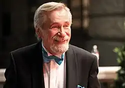 Älterer Mann mit weißem Haar und Bart, der einen schwarzen Smoking und eine blaue Fliege trägt, lächelt freundlich. Der Hintergrund ist unscharf und schafft eine elegante Atmosphäre.