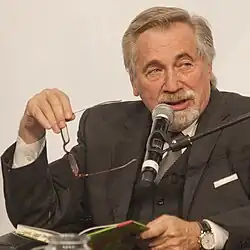 Älterer Mann im Anzug mit Brille, der mit nachdenklicher Miene in ein Mikrofon spricht und ein offenes Buch im Schoß hält.