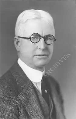 Mann mittleren Alters im dunklen Anzug und weißem Hemd ab Brustkorb im Seitenprofil und in die Kamera schauend in schwarzweiß.