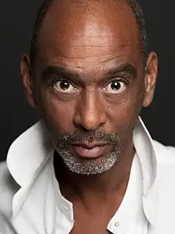 Pierre Sanoussi-Bliss mit kurzen, grauen Bart und intensivem Blick. Er trägt ein offenes, weißes Hemd.Der Hintergrund des Bildes ist dunkel und bildet einen Kontrast zum hellen Hemd.