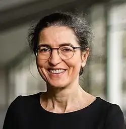 Porträt von Brigitte Röder. Sie ist ca. Mitte 50, trägt die graumelierten dunklen Haare im Dutt nach hinten, eine Brille und ein schwarzes Shirt. Sie lacht frontal in die Kamera.