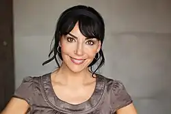 Maria Sanchez auf dieser Oberkörperansicht lächelt geradeaus in die Kamera. Sie ist sehr schlank und trägt ihre glatten schwarzen Haare mit einem gescheitelten Pony zu einem Zopf nach hinten gebunden, dazu ein schwarzes Haarband, das glatt auf dem Kopf anliegt. Ein paar Haarsträhnen sind kunstvoll gelöst und liegen locker hinter ihren Ohren. Sie trägt dünne mittelgroße silberne Kreolen in den Ohren. Ihr T-Shirt ist aus einem glänzend braunen Material und hat einen runden, verstärkten Halsausschnitt.