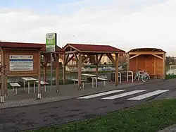 Gruppe von drei Holzdächern und mehreren Schildern ein einem schmalen, asphaltierten Weg. Unter zwei der Überdachungen sind einfache Bänke und ein Tisch aufgestellt, die hinterste hat eine Rückwand aus Holz, an der eine Bank angebracht ist. Zwischen den Überdachungen sind Anlehnbügel für Fahrräder aufgestellt. Im Hintergrund kann man einen Fluss und mehrere hell gestrichene kleinstädtische Häuser erkennen.