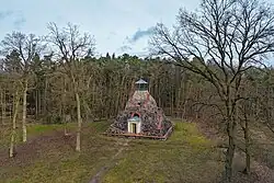 Eine vierseitige Natursteinpyramide mit einer überdachten Spitze und einem hellen Eingangstor steht auf einer Waldlichtung, umgeben von Mischwald.