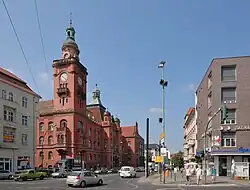 Farbfotografie mit Blick auf eine vierspurige Straße, die zu einem großen roten Gebäude mit auffälligem Uhrenturm führt.