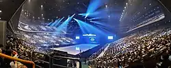 Panoramaansicht des Inneren der voll besetzten Saitama Super Arena zu Beginn der Eisshow „RE_PRAY“, in der Mitte die angestrahlte Eisfläche, im Hintergrund eine Großleinwand mit dem Logo der Show.