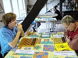 In einem wohnlich eingerichteten Raum stehen hintereinander aufgereiht zwei quadratische Tische mit bunt gemusterten Tischdecken. An beiden Tischen sitzen jeweils links und rechts zwei Schachspieler. Die vorderen beiden sind durch neben ihnen liegende, mit ihren Namen beschriftete Zettel als Dieter Riegler aus Heidelberg (links) und Olaf Dobierzin aus Leipzig (rechts) identifizierbar. Riegler – mit geschlossenen Augen – ist blind, Dobierzin – mit Brille – stark sehbehindert. Hinten auf dem Tisch stehen eine Flasche Mineralwasser, ein Holzkästchen für die Schachfiguren (darin ein weißes Stofftierchen – möglicherweise Rieglers Maskottchen) und eine moderne Schachuhr mit Digitalanzeige. Vor jedem Spieler steht ein eigenes Blindenschachbrett. Riegler hat mit d2–d4 eröffnet, beide Bretter zeigen diese Stellung an. Riegler hat sich abwartend zurückgelehnt, Doberzin konzentriert sich über sein Brett gebeugt auf den Gegenzug.