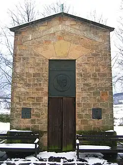 Portalseite des Sprudelhauses mit Holztür und zwei Ruhebänken