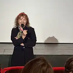 Die französische Schauspielerin Rosette steht auf einer Bühne und hält ein Mikrofon in der rechten Hand. Sie trägt eine dunkle Jacke und einen Schal. Hinter ihr ist eine große weiße Leinwand sichtbar, die den Hintergrund der Bühne bildet. Im Vordergrund des Bildes sitzen einige Menschen, die in Richtung der Bühne blicken. Die Umgebung lässt vermuten, dass es sich um einen Vortrag, eine Präsentation oder eine Aufführung in einem Theater oder Auditorium handelt.