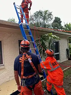 Zwei Männer in mit SES beschrifteter Warnkleidung stehen vor einem einstöckigen Haus und halten eine an dieses gelehnte Leiter. Auf dem Dach steht ein weiterer Mann mit einem Seil.