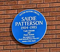 Das in einem leichten Winkeln aufgenommene Farbbild zeigt mittig die runde, blaue Plakette auf einer sonst gleichmäßigen Wand roter Backsteinziegel mit dunkelgrauem Mörtel. Due Aufschrift auf der Plakette lautet: „Ulster History Circle — Saidie Patterson — 1904–1985 — Trade Unionist — and — Peace Activist — first recepient of the — World Methodist Peace Price 1977 — member of this church“