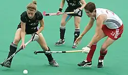 Sally Walton im weißen Trikot und mit rotem Rock bedrängt die ballführende deutsche Spielerin.