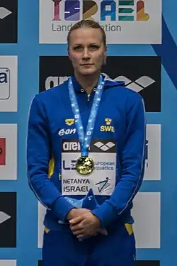 Sarah Sjöström bei einer Siegerehrung im blauen Trainingsanzug mit drei gelben schwedischen Kronen. Um den Hals am hellblauen Band trägt Sjöström eine Goldmedaille.