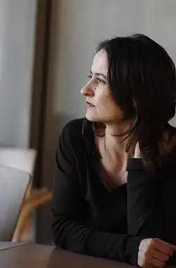 Sasha Waltz sitzt an einem Tisch und schaut nachdenklich zur Seite. Sie hat schulterlanges, dunkles Haar und trägt ein langärmliges, dunkles Oberteil. Die linke Hand ruht entspannt auf dem Tisch. Die rechte Hand berührt die Seite des Halses, was eine nachdenkliche oder entspannte Haltung andeuten könnte.Der Hintergrund scheint eine Innenraumszene zu zeigen. Es sind ein Stuhl und eine Wand zu erkennen, die schlicht gehalten sind und nicht stark vom Hauptmotiv ablenken.