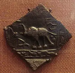 Eine quadratische Münze aus dunklem Metall, eine Ecke zeigt nach unten. Darauf ist ein Elefant mit erhobenem Rüssel dargestellt