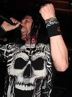 Großaufnahme des langhaarigen Sängers mit Mikrofon in der rechten Hand, zum Singen geöffnetem Mund und geballter linken Faust