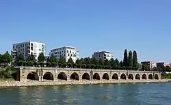 Teil einer Betonkonstruktion am Rheinufer mit 14 Rundbögen, über die ein Geh- und Radweg führt. Dahinter weiße, kubische Wohnhäuser