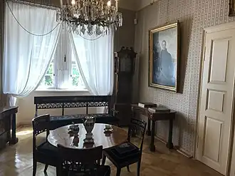 Farbige Eckansicht eines Innenraums mit einem runden Tisch, einem Kaffeeservice und drei Stühlen in der Mitte. An der linken Wand sind Fenster mit weißen Gardinen und einer verzierten Bank davor. Rechts vom Fenster befindet sich eine Standuhr. An der rechten Wand mit braun-weißem Muster hängt ein Porträt, unter dem ein Schreibtisch mit Büchern steht. Rechts davon eine weiße, verschlossene Tür. Am oberen Bildrand strahlt ein Kronleuchter.