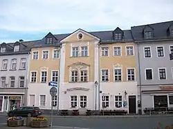 Häuserreihe hinter einer Straße ohne Fahrbahnmarkierung. Das mittlere Haus in gelb und weiß sieht ordentlich aus, kein Neubau. Einbahnstraßenschild zeigt Richtung Betrachter*in und weg von der Straße, deutet also eine Kreuzung an.