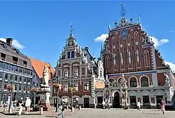 Frontale Farbfotografie von zwei weiß-braunen Gebäuden mit verzierten Staffelgiebeln. An jeder Staffelecke stehen Figuren und kleine Obelisken. An den Dachspitzen befinden sich Eisenkreuze. An der rechten Fassade hängen vier Statuen und vor dem rechten Gebäude ist ein flacher Vorbau mit einem verzierten Torbogen, in dessen Pfeilern zwei Farbfiguren stehen. Auf dem Vorplatz befindet sich eine weiße Ritterstatue mit Wappenschild.