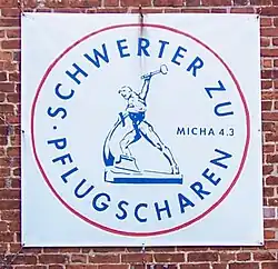 An einer roten Ziegelwand hängt ein Banner mit dem kreisrunden Symbol der Friedensbewegung „Schwerter zu Pflugscharen“. Es ist rot umrandet. Im Kreis steht oben entlang des Randes „Schwerter zu“ und unten „Pflugscharen“. In der Mitte ist die gleichnamige Bronzeskulptur abgebilder.