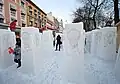 Frontale Farbfotografie von vier Reihen mehrerer Eisskulpturen, die in Rechteckform an allen vier Seiten den schreienden Menschen eingeschnitzt haben. Menschen laufen durch und im linken Hintergrund ist eine Häuserfassade zu sehen.