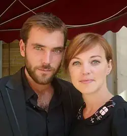 Matteo van der Grijn und Loes Haverkort, die eng beieinander stehen. Er trägt ein schwarzes Jackett über einem schwarzen Hemd, dessen oberster Knopf geöffnet ist. Sie ist in einem schwarzen Kleid gekleidet, das am Kragen mit dekorativen Elementen versehen ist. Diese dekorativen Elemente sind glänzend und in einem Muster angeordnet. Im Hintergrund ist ein rotes Vordach und eine Backsteinwand zu sehen, was möglicherweise auf einen städtischen oder architektonischen Kontext hindeutet. Die Darstellung wirkt elegant und zeitgenössisch.