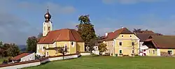 Spätbarocke Pfarrkirche mit Zwiebelturm, rotem Ziegeldach und gelbem Verputz; rechts daneben in gleicher Farbgebung der Pfarrhof mit Krüppelwalmdächern