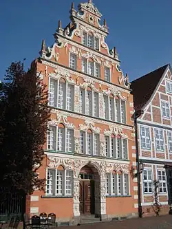 Seitliche Farbfotografie eines dreigeschossigen, weiß-braunen Gebäudes mit Staffelgiebel, an deren Ecken kleine Obelisken stehen. Die Fenster und der Torbogeneingang sind mit weißen Ornamenten versehen. Rechts steht ein Haus mit weißen Zierbalken im Bauernhausstil.