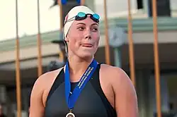 Elaine Breeden im dunklen Badeanzug mit weißer Badekappe trägt um den Hals ein blaues Band mit einer Medaille.
