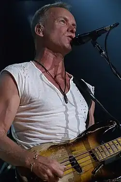 A man with a v-neck, white T-shirt wearing a necklace and bracelet standing behind a microphone, holding a guitar.