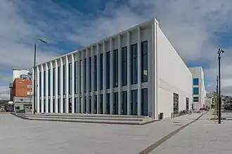 Foto eines weißen Gebäudes, auf der Vorderseite viele lange schmale Fenster, auf der Seite hingegen kaum Fenster