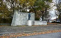 Das Ensemble des Ernst-Thälmann-Denkmals in Stralsund im Jahr 2023 mit einem weiten Platz, einer niedrigen begrenzenden Mauer, dem auf einem Sockel stehenden Standbild Thälmanns und einer nach hinten abgernzenden hohen Sandsteinmauer.