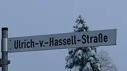 Foto eines weißen Straßenschildes vor grauem Winterhimmel mit der schwarzen Aufschrift Ulrich-v.-Hassel-Straße