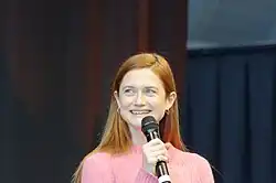 Frontale Farbfotografie von einer jungen, rothaarigen Frau, die lächelnd nach links blickt. Sie hält in ihrer rechten Hand ein Mikrofon. Ihre langen, glatten Haare fallen hinter ihren Rücken und sie trägt einen pinken Pullover. Im Hintergrund sind dunkle Vorhänge zu sehen.