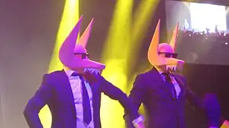 Foto zweier Personen, die mit einer gelben Maske und Sonnenbrille auf einer Bühne stehen