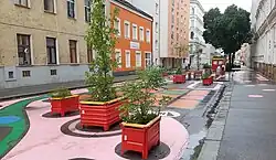 Mit bunten Mustern bemalte Straße vor einer Schule, die aufgrund von jungen Baumpflanzungen und Stadtmöbeln nur noch den Weg für Fußgehende und Radfahrende erlaubt