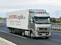 Photo eines auf einer mehrspurigen Straße fahrenden Sattelzuges, an dessen Sattelzugmaschine vorne eine Plakette mit der Aufschrift „V“ für „EURO V sicheres“ Fahrzeug angebracht ist.