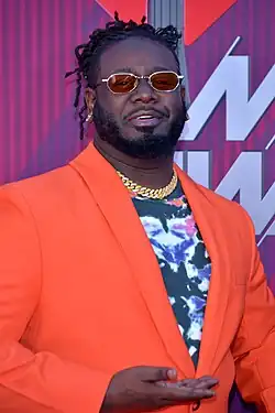 T-Pain steht vor einer lila Wand mit weißer Aufschrift, die verdeckt ist. Er trägt ein orangenes Sakko, ein schwarzes Hemd mit Blumenaufdruck, eine goldfarbene Halskette, einen Dreitagebart, eine Sonnenbrille mit roten Gläsern und die schwarzen Haare als kurze Dreadlocks.