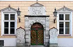 Frontale Farbfotografie vor dem Eingang einer orangen Fassade. Die Tür ist mit roten Querstreifen und goldenen Blumen verziert. Im oberen Teil ist eine Figur in einem Wappen. Um die Tür sind mit grauem Stein Ornamente eingemeißelt, die auch in einem Quadrat und einem Giebelfeld über der Tür erscheinen. Im Quadrat halten zwei Löwen ein Wappen. An beiden Seiten der Tür stehen zwei gemeißelte Steinblöcke und zwei Laternen hängen zwischen Tür und zwei Fenstern.