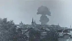 Schwarzweißfotografie von einer Stadt mit einer riesigen Rauchwolke. Einige Kirchtürme ragen hervor und vorne bedecken Bäume manche Häuser.